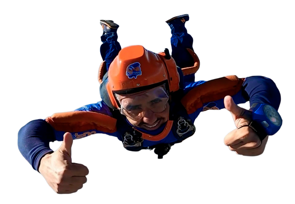 Um paraquedista sorridente, vestindo um macacão azul e laranja e capacete laranja, realiza um salto livre com os polegares levantados, flutuando no ar contra um fundo transparente